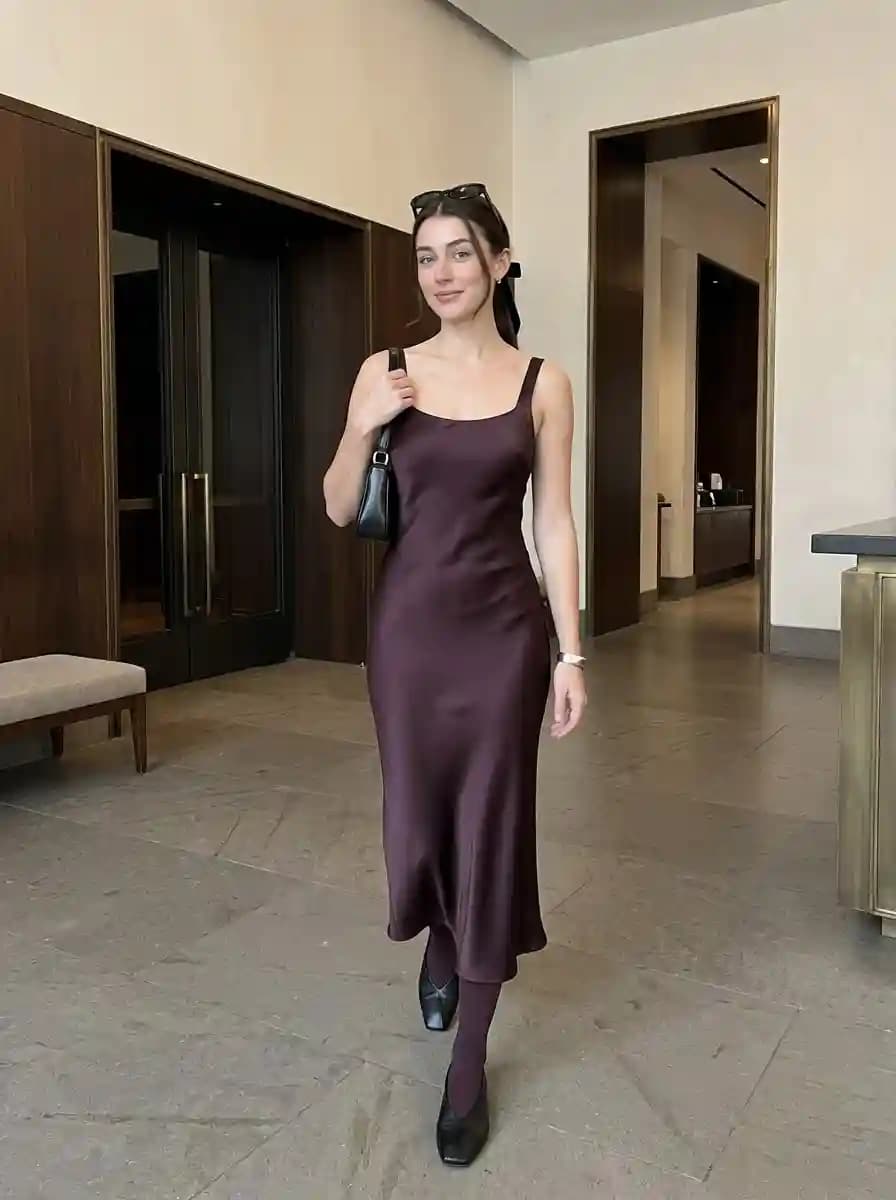 Woman wearing a deep burgundy satin midi slip dress with dark sheer tights, black pumps, and a small black shoulder bag.